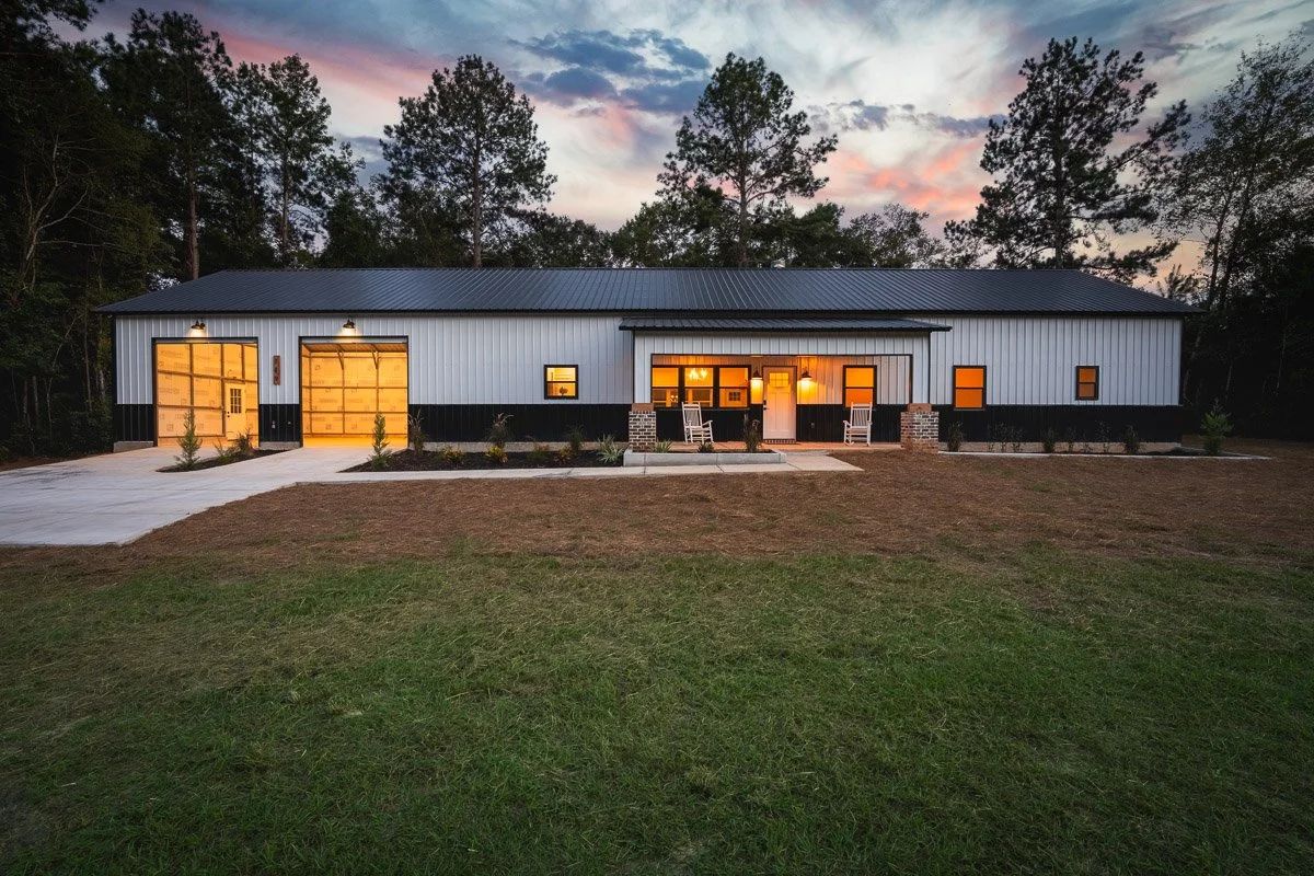 Real Estate photo of a modern barndominium-style home with metal siding, lit from inside, surrounded by trees, with a small front porch and two rocking chairs, located in a rural area at sunset.