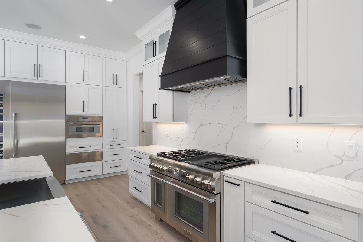 Modern white kitchen with marble countertops, black cabinet handles, stainless steel appliances, and a black range hood.