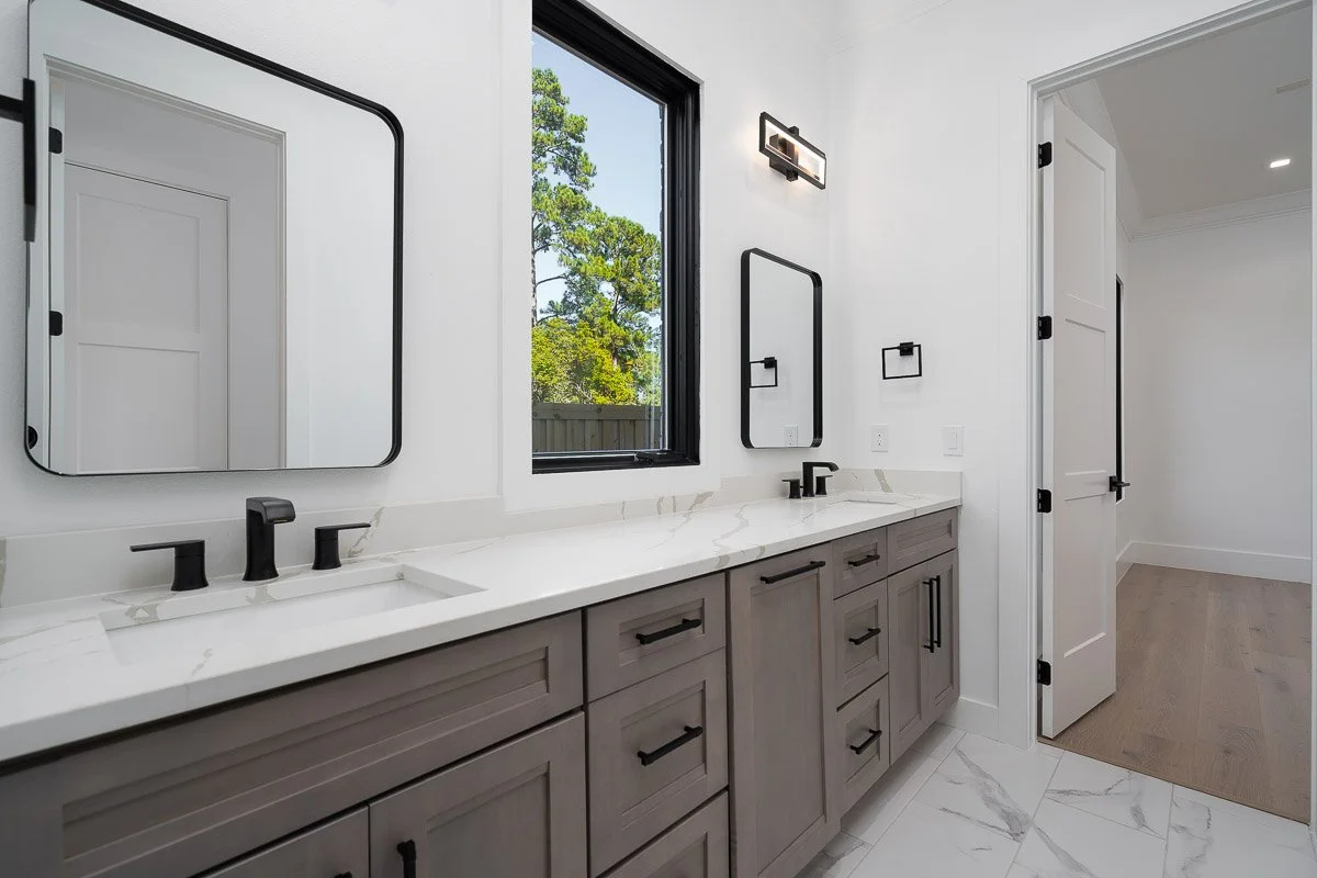 Modern bathroom with double vanity, white marble countertop, black fixtures, two mirrors, and the window showing greenery outside.