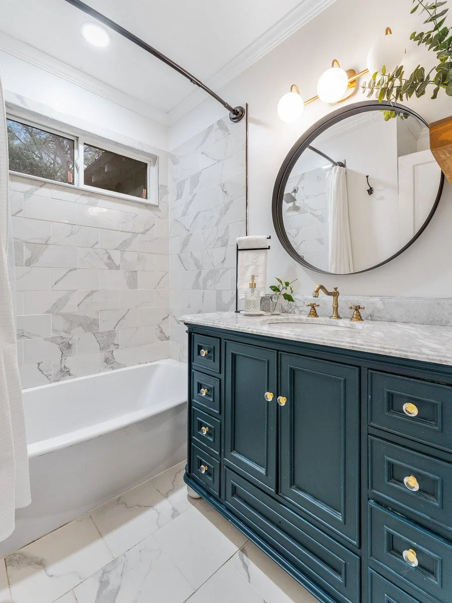 Modern bathroom with white marble tiles, a navy blue vanity with gold knobs, round mirror, wall light fixture, and a small window near the ceiling.