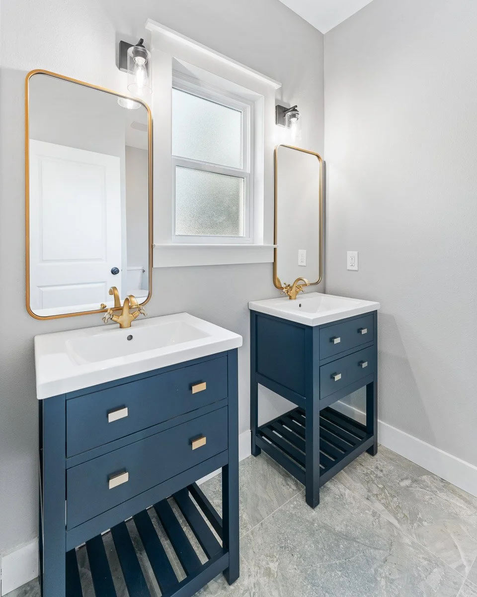 Real Estate photo of a two navy blue bathroom vanities with white sinks, gold fixtures, rectangular mirrors with gold frames, a window, and modern wall sconces in a bright bathroom with light gray walls and gray tiled flooring.
