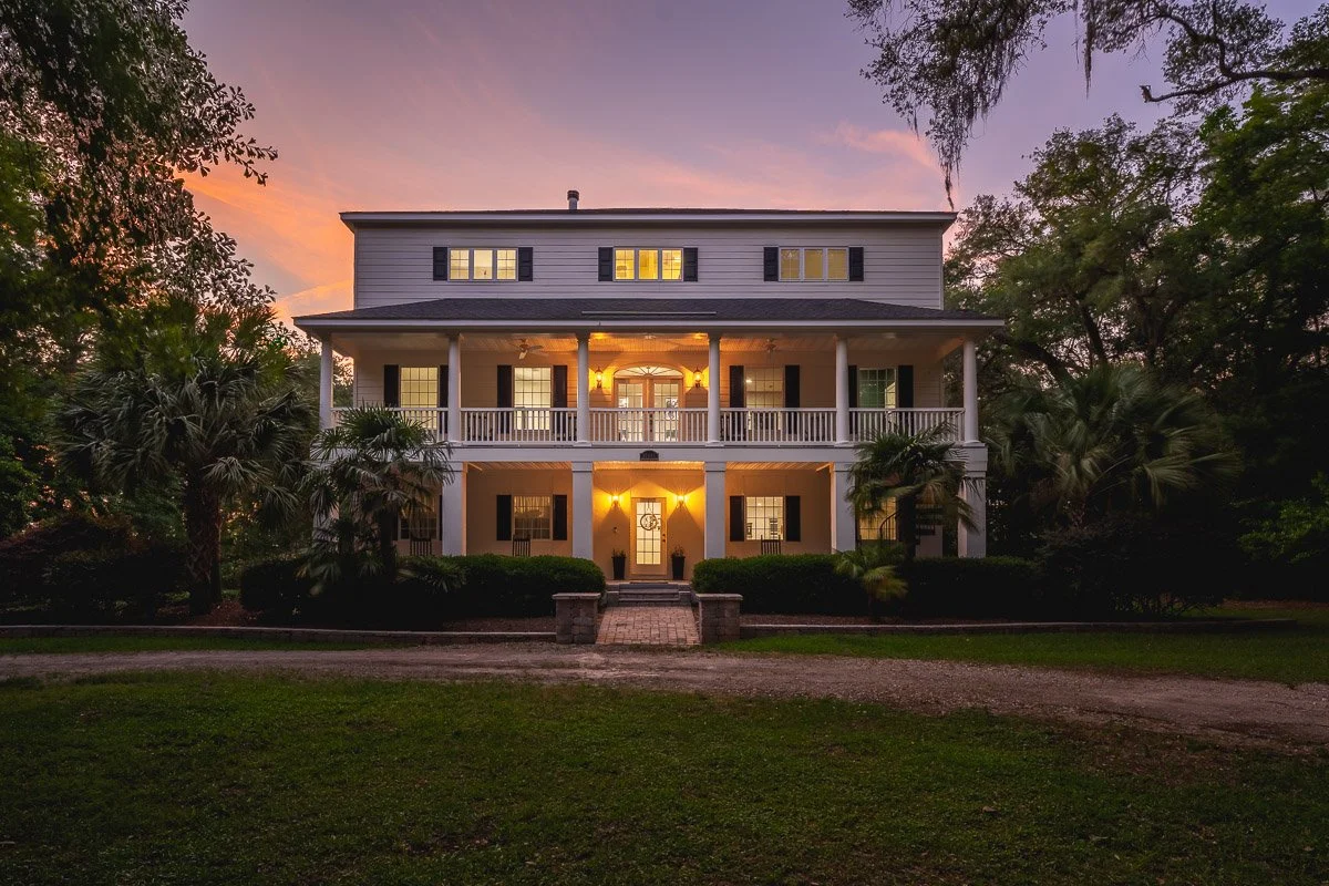 Real Estate photo of a large, plantation style home with a lit porch and windows, surrounded by palm trees and greenery, at sunset.