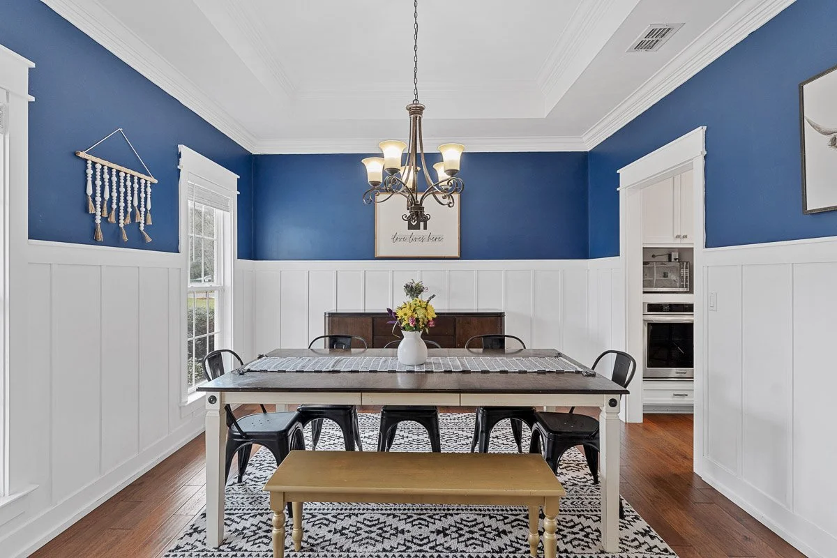 A dining room with white wainscoting and blue walls, a wooden table with a patterned table runner, black chairs, a white vase with flowers, a chandelier hanging from the ceiling, a wall art with the text 'love lives here', and a window with white bli