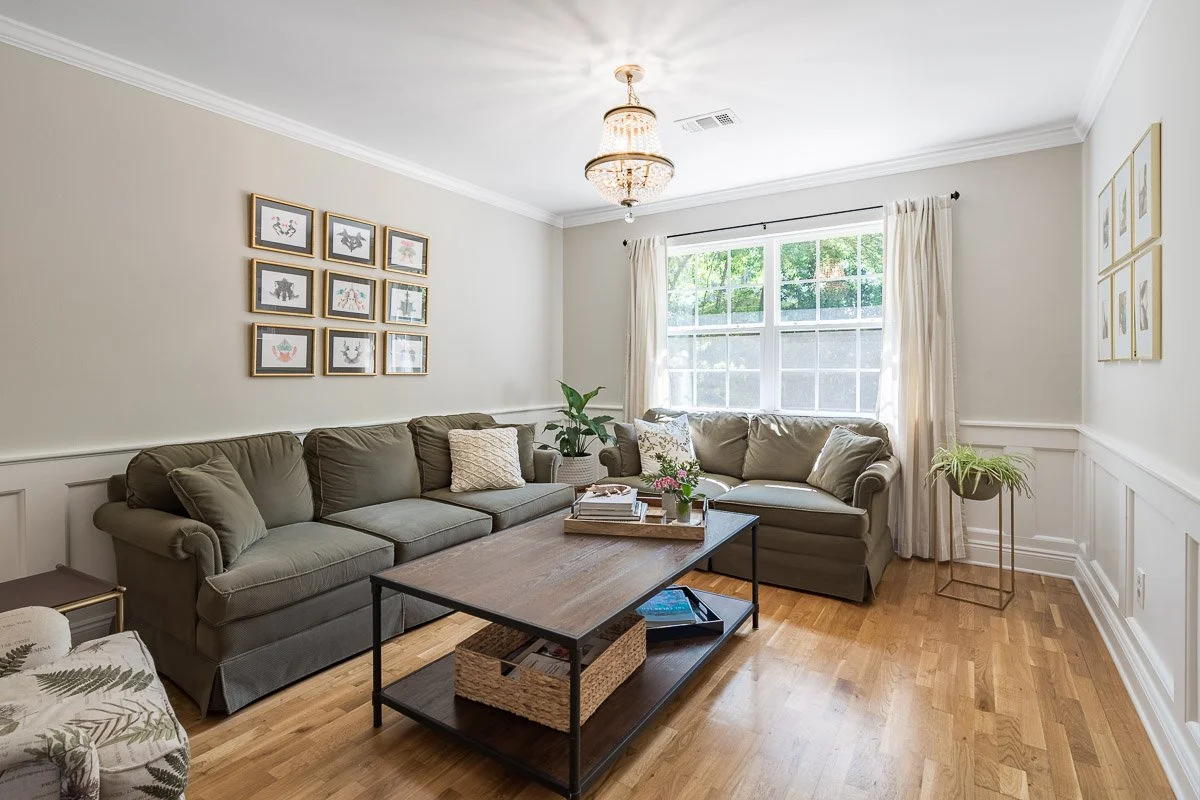 Real Estate photo of a living room with warm beige walls, hardwood floors, two green sofas, a wooden coffee table with books and plants, a chandelier, window curtains, and framed art on the walls.