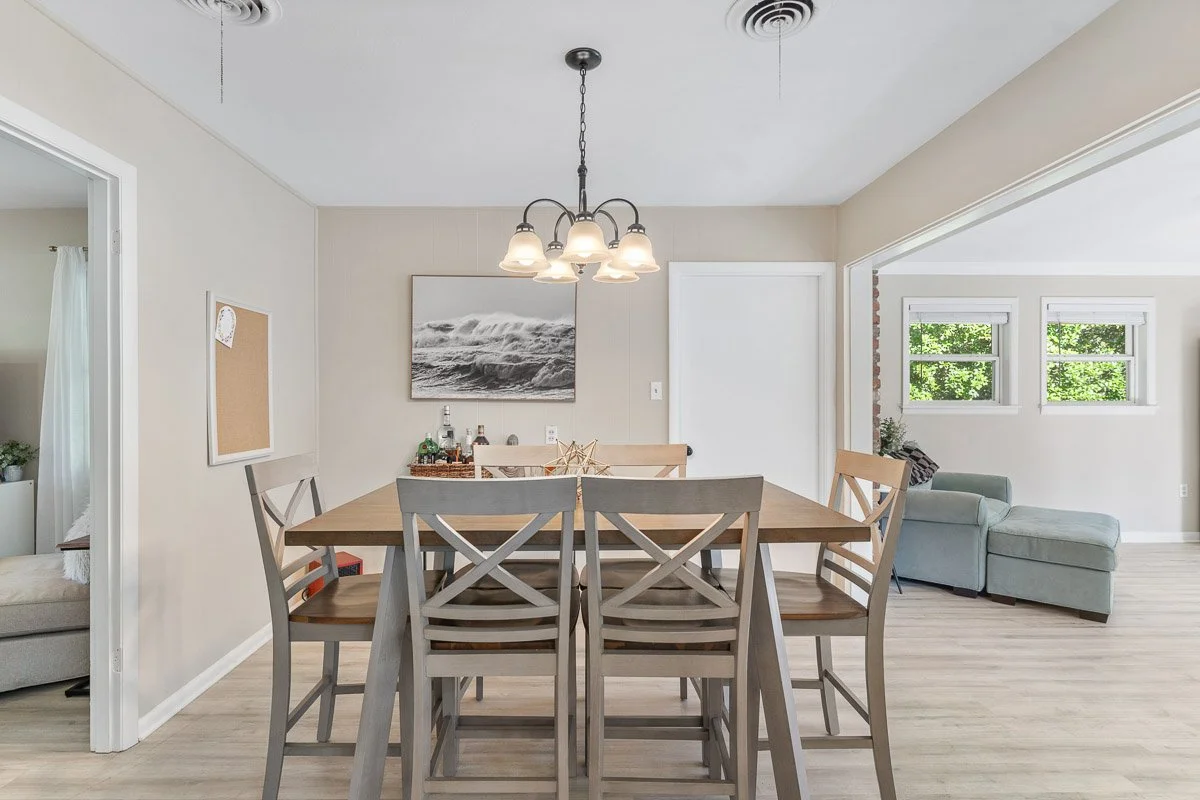 Dining room with a wooden table and six chairs, chandelier overhead, wall art, and a living area with a light green sectional sofa and windows