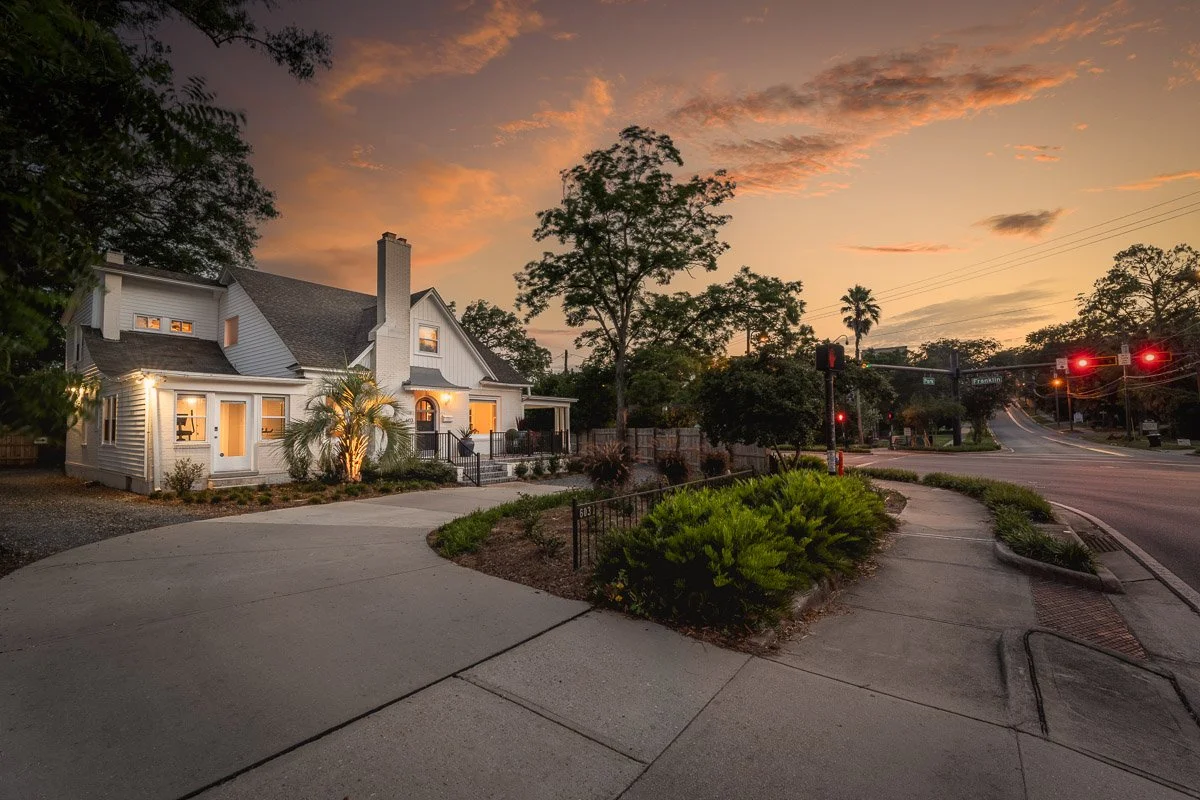 Real Estate photo of a cozy white house with lights on, next to a street corner during sunset, with a cloudy sky, trees, and streetlights.