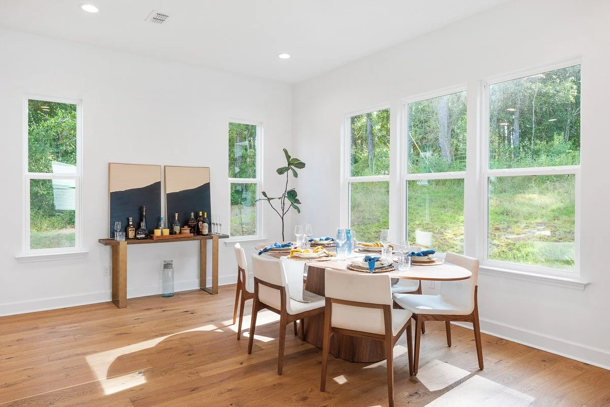 Real Estate photo of a bright dining room with white walls, hardwood floors, and large windows showing a green outdoor area. There is a round wooden table set for a meal with six chairs, and a small wooden side table.