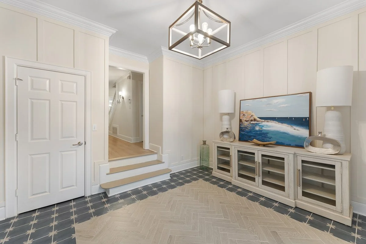 Real Estate photo of an entryway with white walls, a white door, a beige and black tiled floor, and a white wooden console table holding a landscape painting of a seascape, two white lamps, and decorative objects.