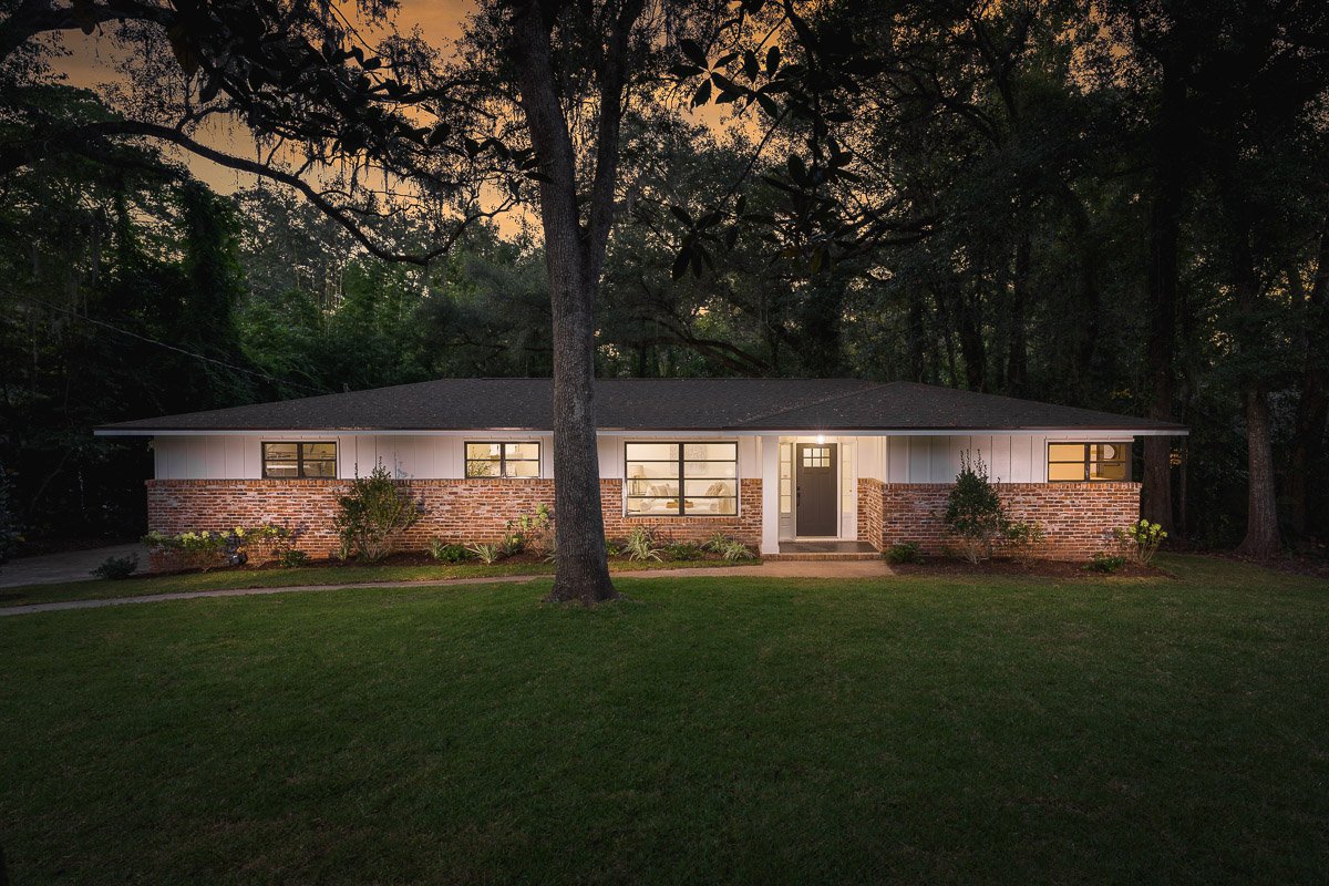 A single-story house with a brick and white siding exterior, large front windows, and a central front door illuminated from inside, surrounded by a well-maintained lawn and trees, at dusk.