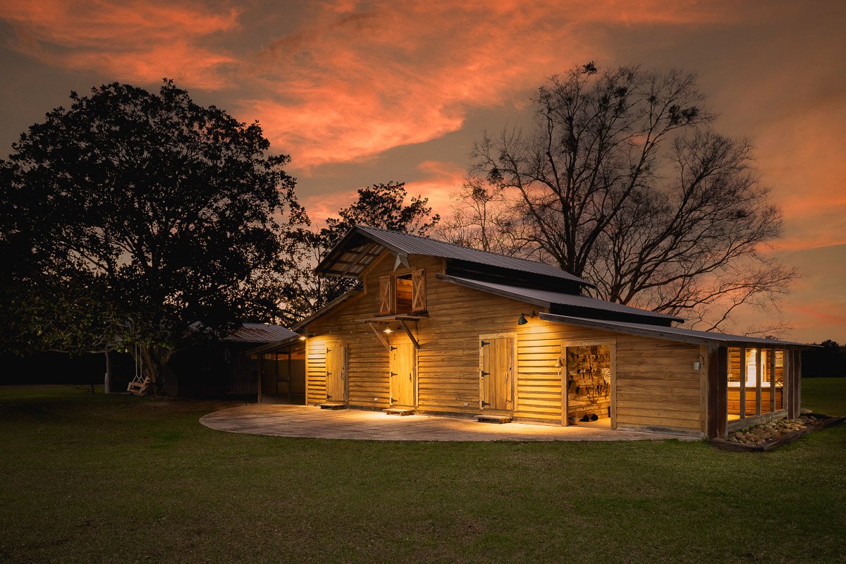 A wooden barn with lights on, set against a sunset sky with wispy clouds and trees in the background.