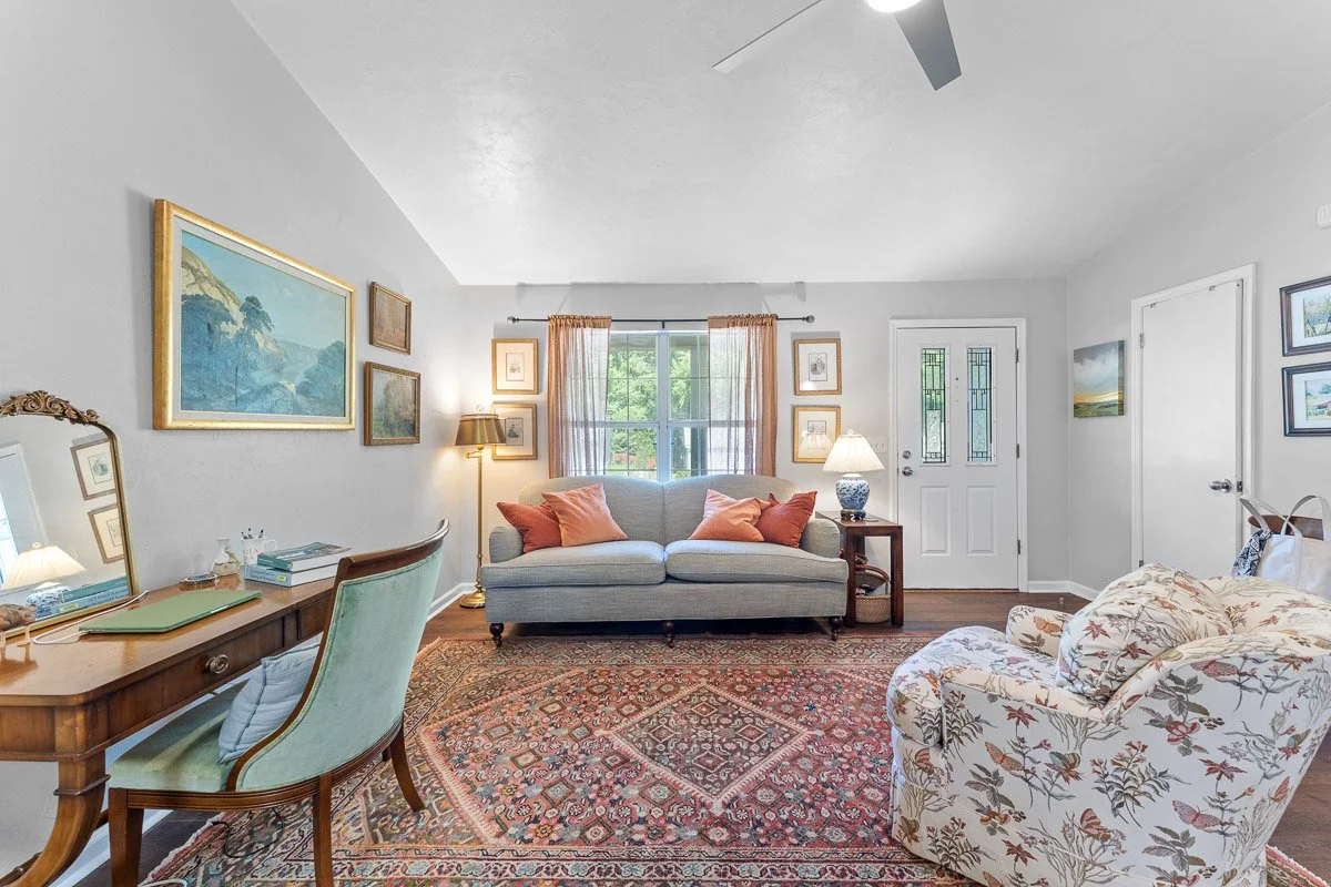 Real Estate photo of a living room with a light gray sofa, floral armchair, wooden desk with mirror, and various wall art, with a window and various lamps providing lighting.