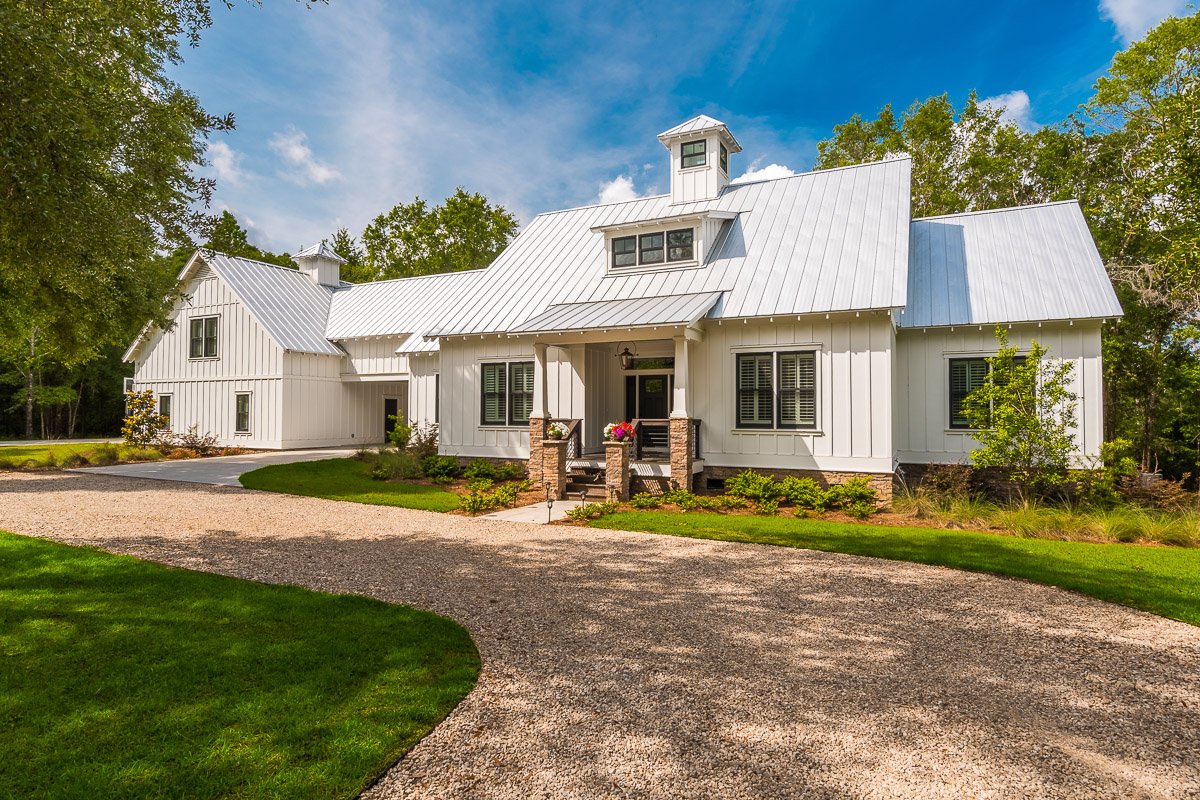 Real Estate photo of a white farmhouse-style home with metal roof, surrounded by green lawn and trees, with a gravel driveway in front.