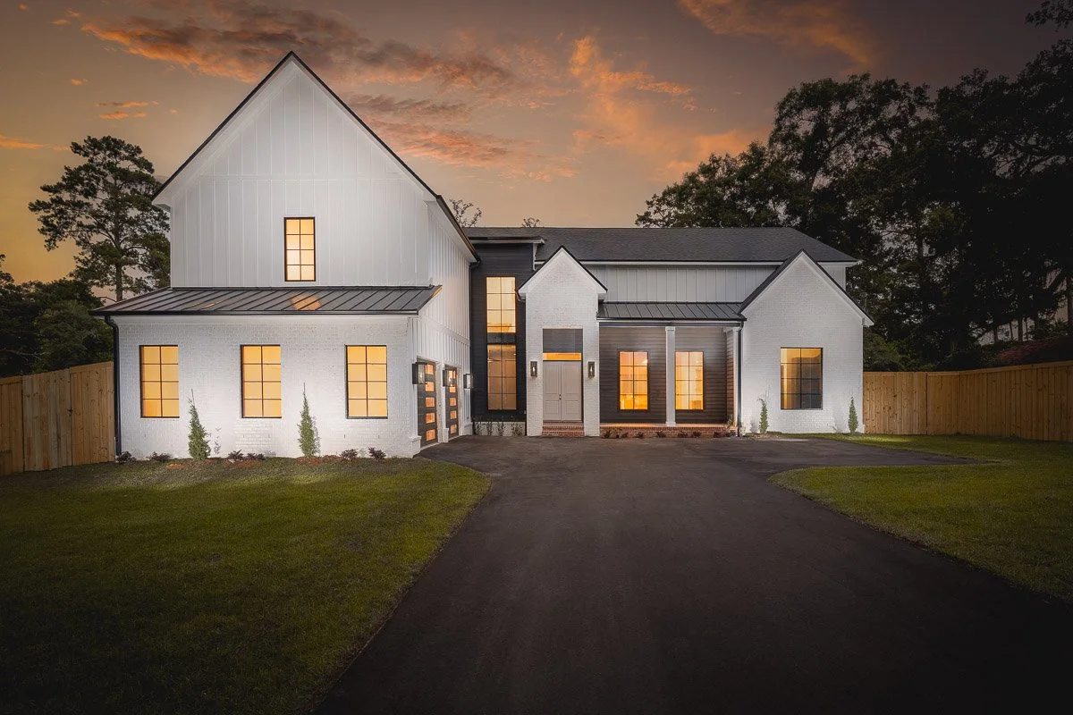 Real Estate photo of a luxury modern two-story home with white exterior walls, large illuminated windows, and a dark metal roof, situated on a well-manicured lawn at dusk.