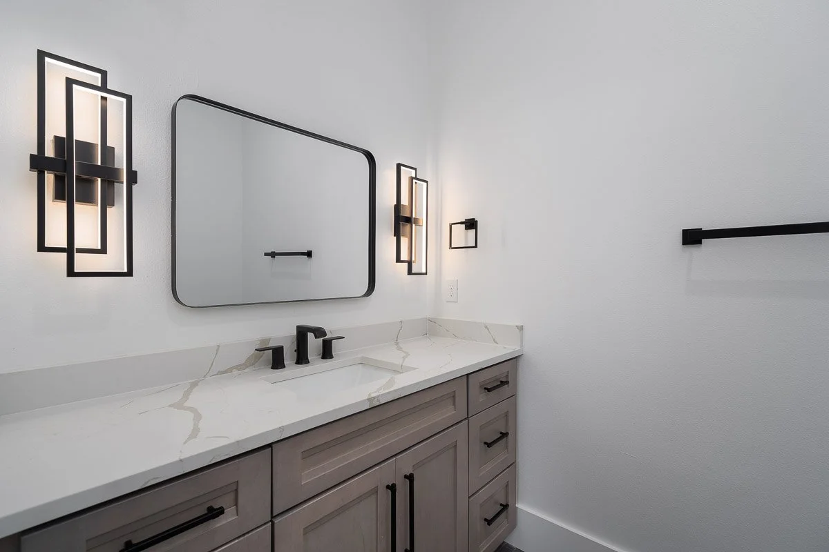 Modern bathroom vanity with gray cabinets, white marble countertop, black faucet, rectangular mirror, and black wall-mounted lights.