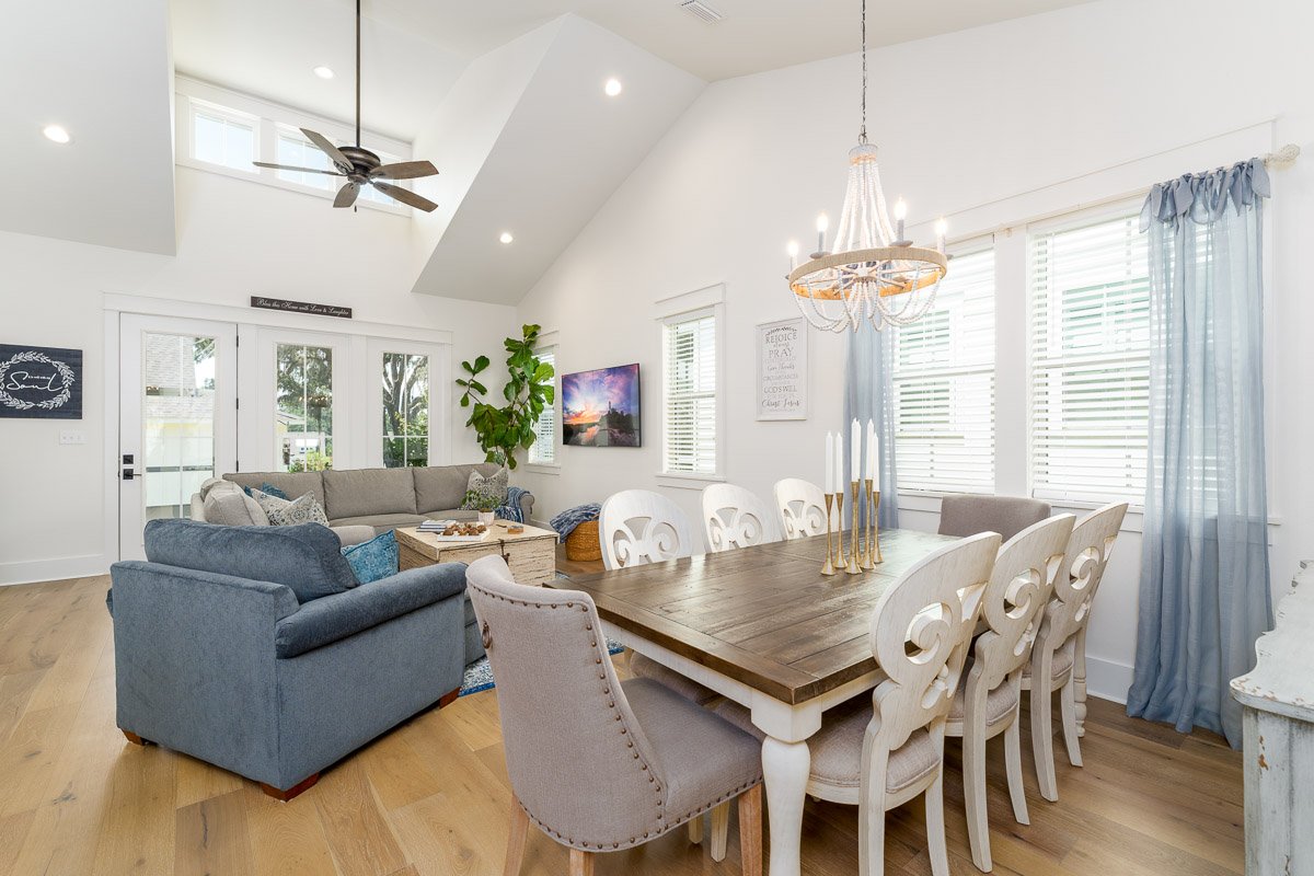 Real Estate photo of a bright living room with white walls, large windows with blue curtains, a chandelier, a wooden dining table with six white chairs, a gray sofa, a blue armchair, a ceiling fan, and a TV on the wall.