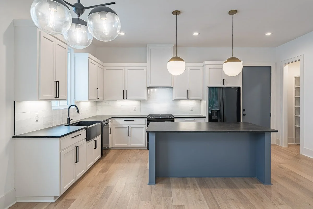Real Estate photo of a modern kitchen with white cabinets, black countertops, a black refrigerator, pendant lights, and a blue island with a dark countertop.