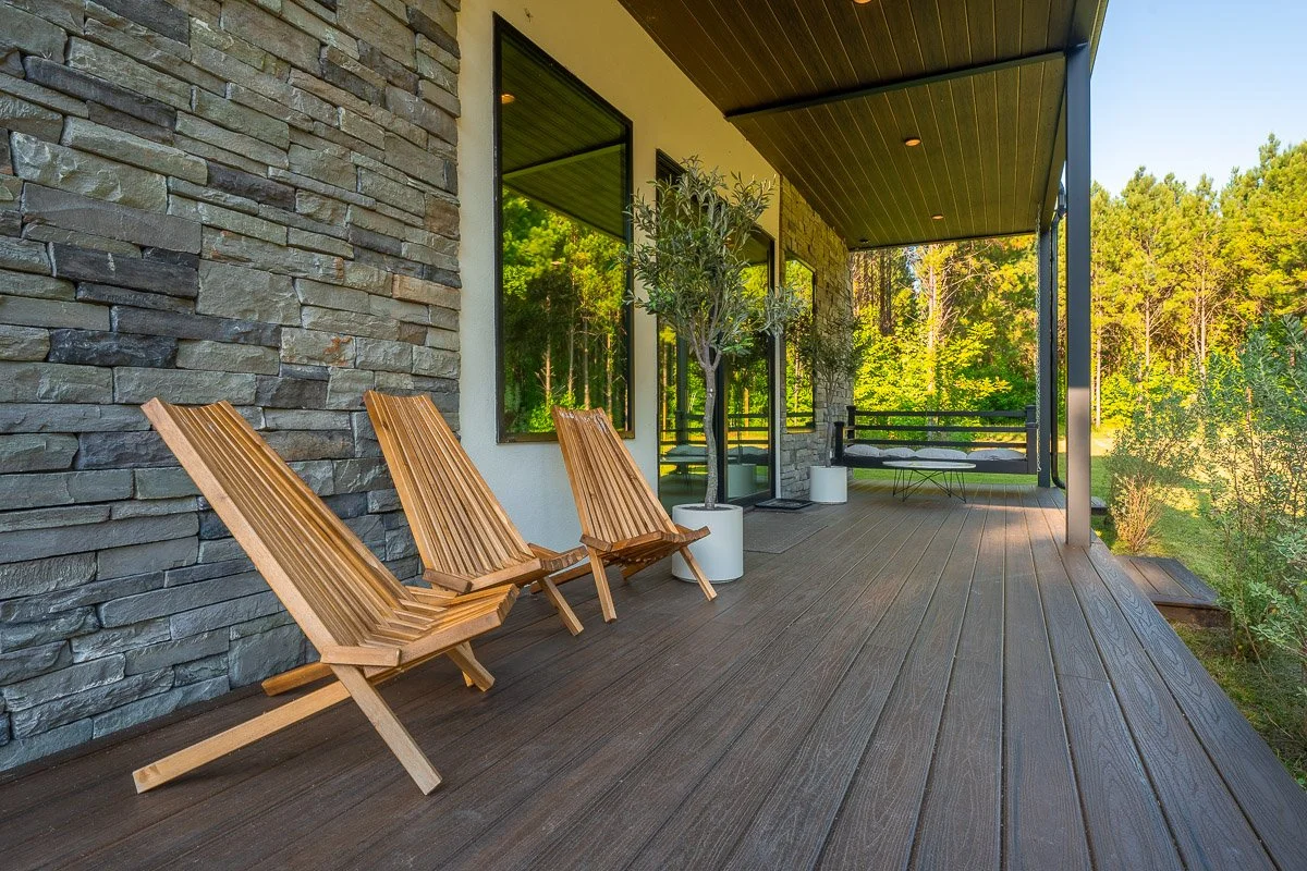 Real Estate photo of a  spacious outdoor porch with wooden decks and outdoor furniture, including three wooden lounge chairs, two potted trees, a black bench, and a small table, surrounded by green trees and a forest in the background.
