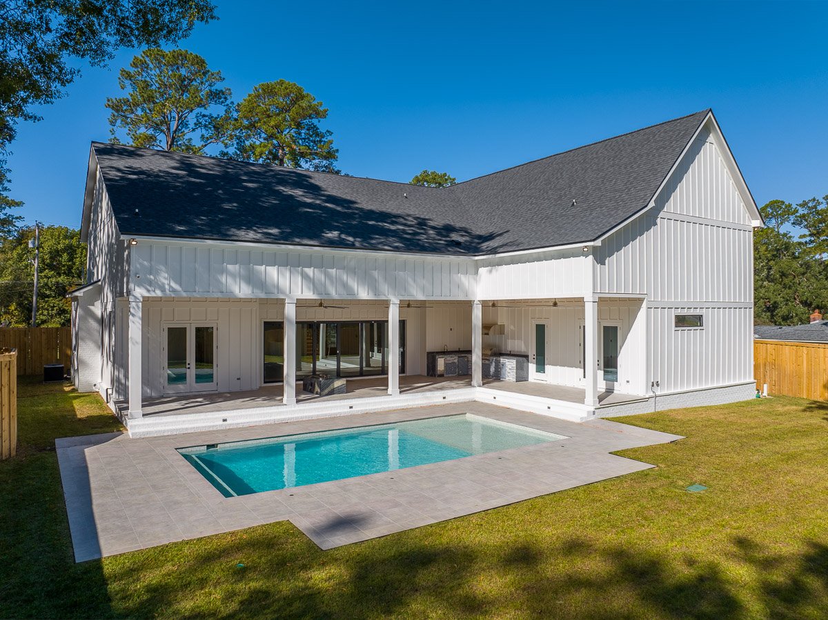 Backyard view of a modern white house with a swimming pool, covered patio, and a grassy yard, under a clear blue sky.