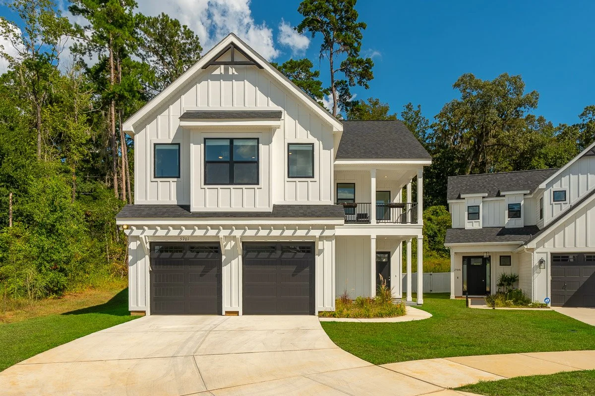 Real Estate photo of a modern white two-story house with black garage doors, black window frames, and a small front porch, surrounded by green trees and grass under a blue sky.