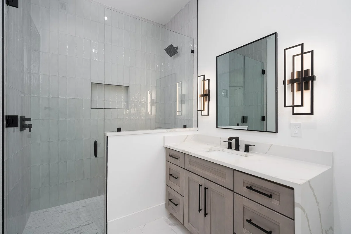 Modern bathroom with white tiled shower area, glass door, grey vanity with black hardware, large mirror, and contemporary black wall sconces.