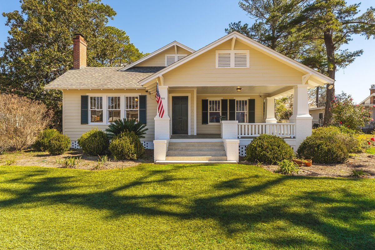Front view of a yellow house with a covered porch, green shutters, steps leading to the front door, and a landscaped yard with bushes and grass.