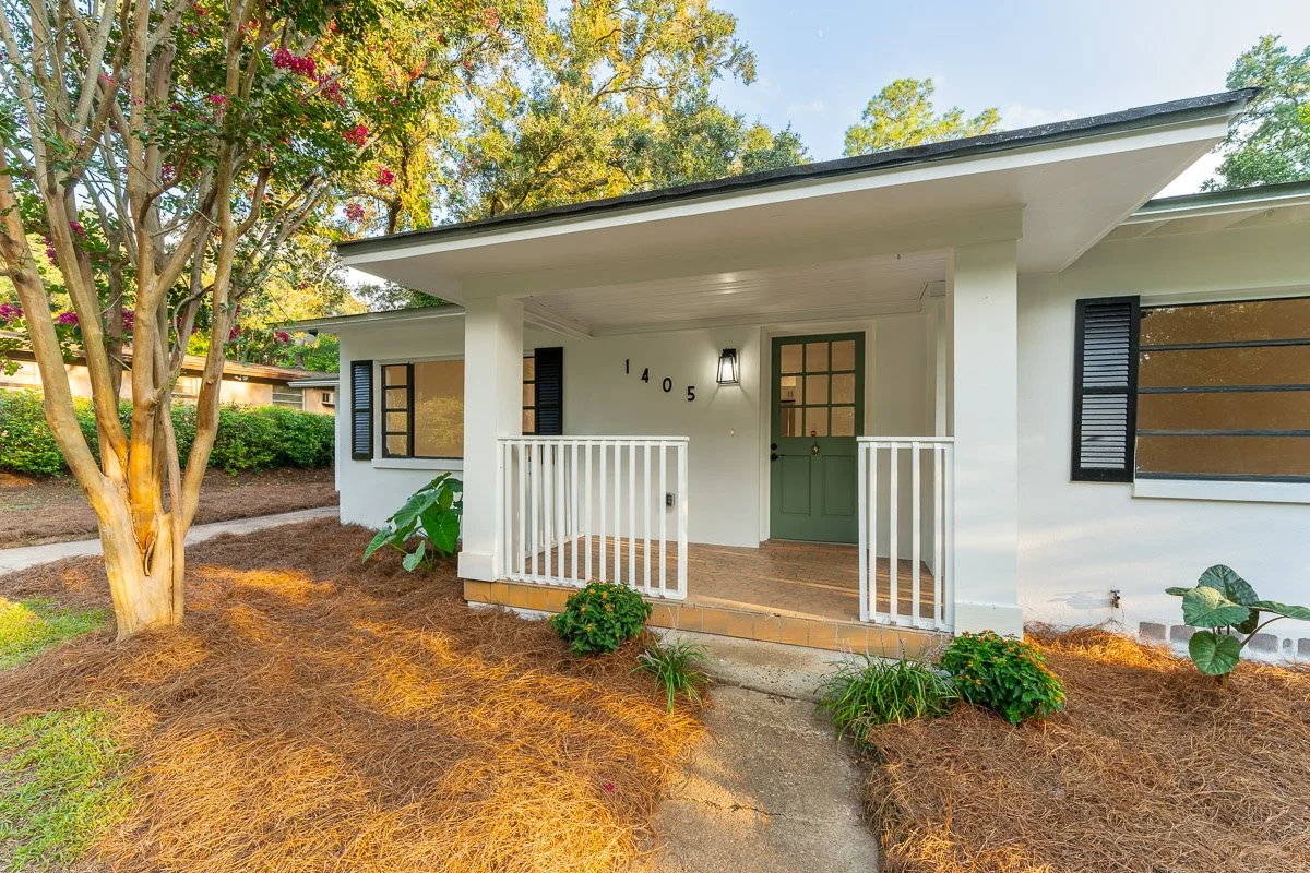 Real Estate photo of a newly renovated home with a terracotta porch, green door, black shutters, and a small garden with bushes and trees.