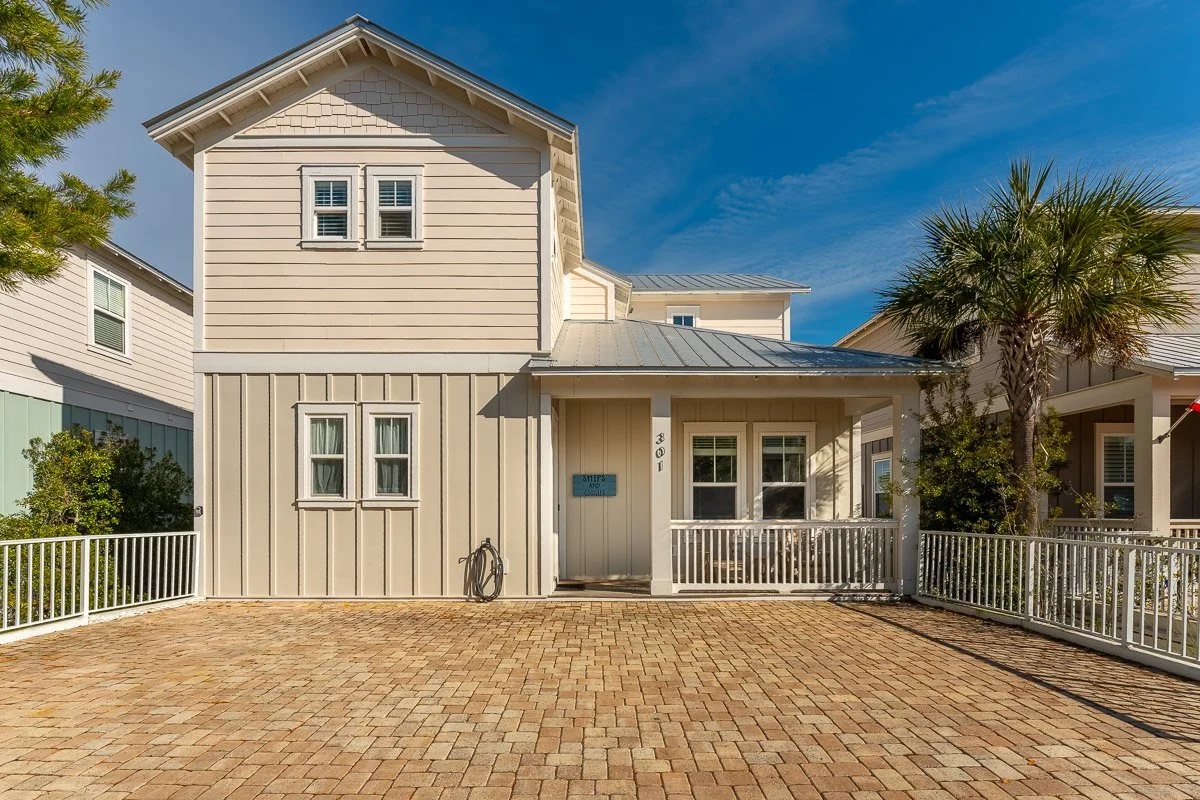 Real Estate photo of a 30A beach house with a brick driveway, white fence, and palm tree, under a blue sky.