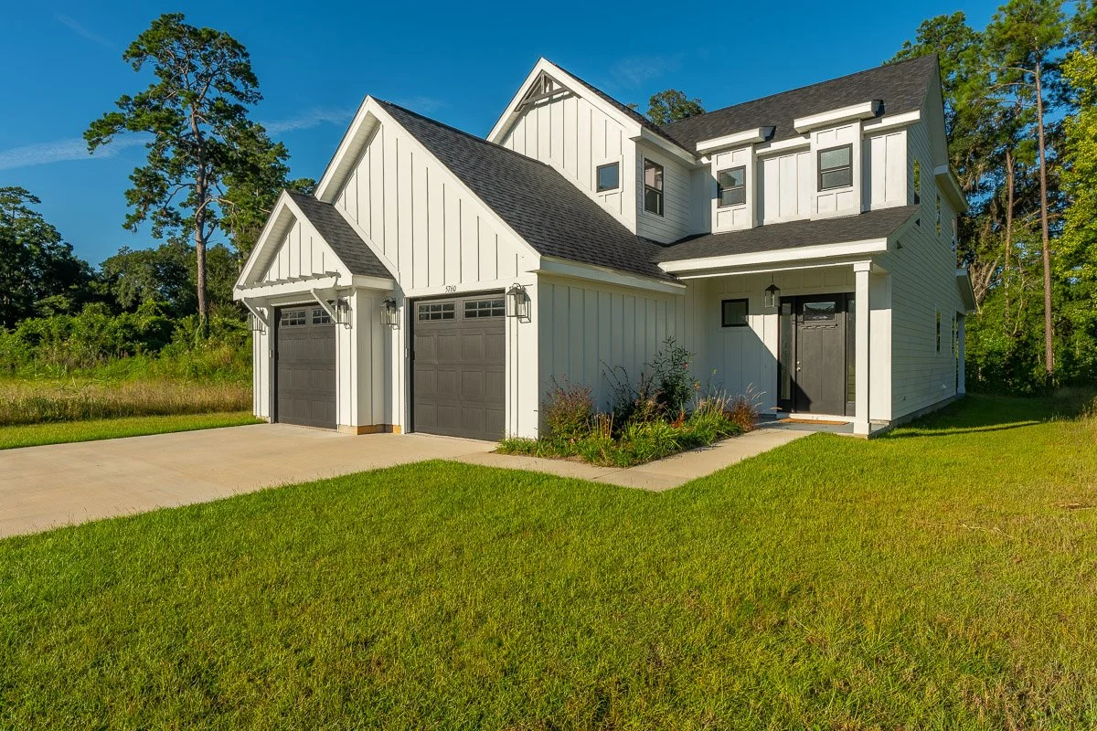 Real Estate photo of a modern two-story house with white exterior walls, black shutters, and a dark gray front door, with attached two-car garage, surrounded by a well-maintained lawn and trees.