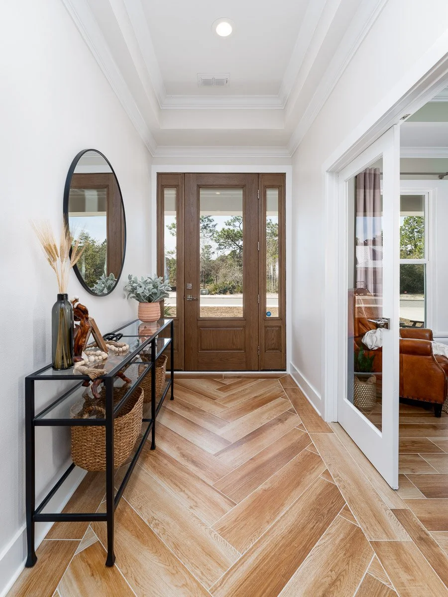 Modern entryway with wooden front door, glass side panels, and a chevron wood floor. Includes a black console table with decorative items, a mirror, and a partially visible room with a leather armchair.
