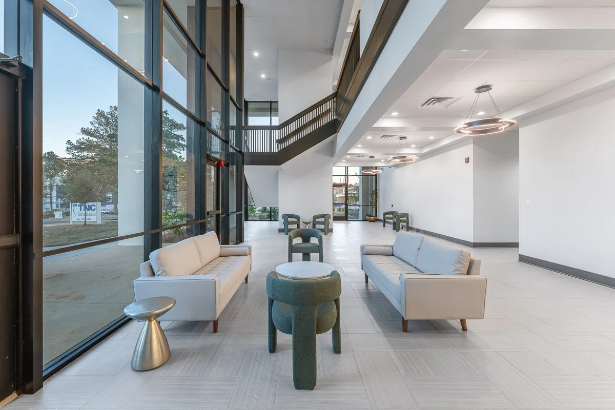 Commercial real estate photo of a modern lobby area with white sofas, green chairs, a round table, and large glass windows with trees outside.