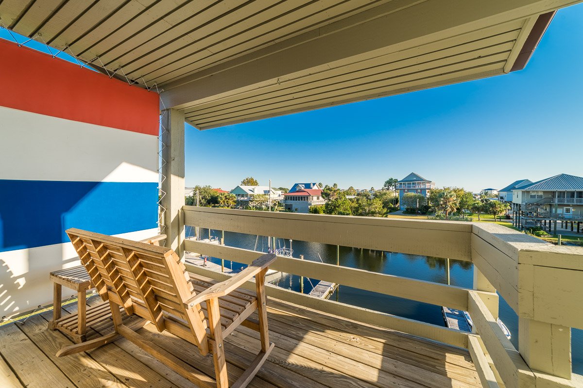 Real Estate photo from the balcony of a waterfront condo, overlooking the canal with houses in the background, decorated with a red, white, and blue flag, and a wooden lounge chair.