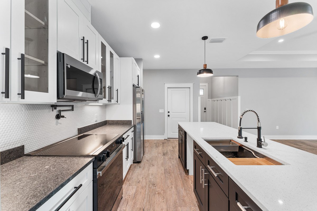 Real Estate photo of a modern kitchen with white upper cabinets, black hardware, stainless steel appliances, a white kitchen island with a black and white faucet, light wood flooring, and pendant lights.