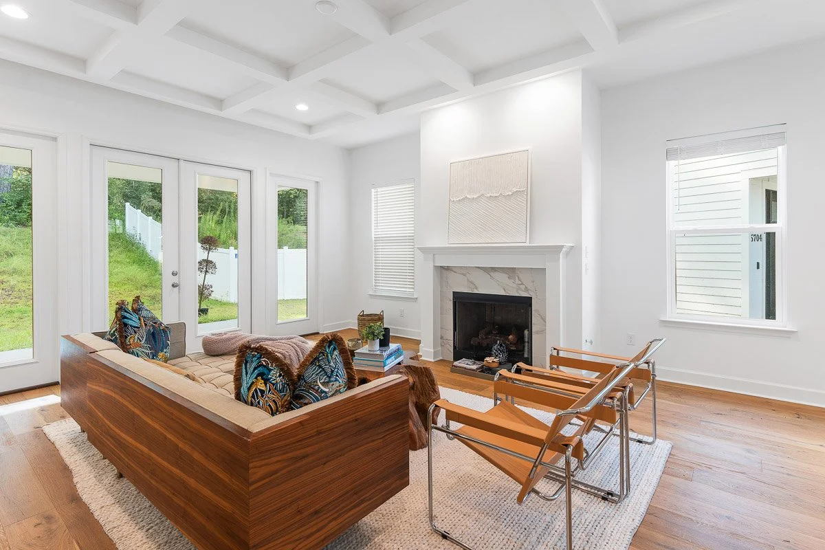Real Estate photo of a living room with white walls, hardwood floors, a white ceiling with recessed lighting, a white fireplace with a marble surround, and large windows and glass doors bringing in natural light.