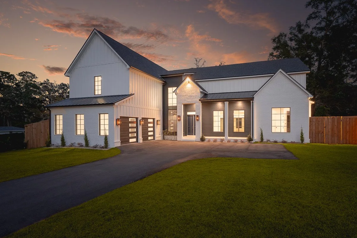 Modern white house with a dark roof, illuminated windows, and a two-car garage at dusk, surrounded by a well-maintained lawn and a wooden fence.