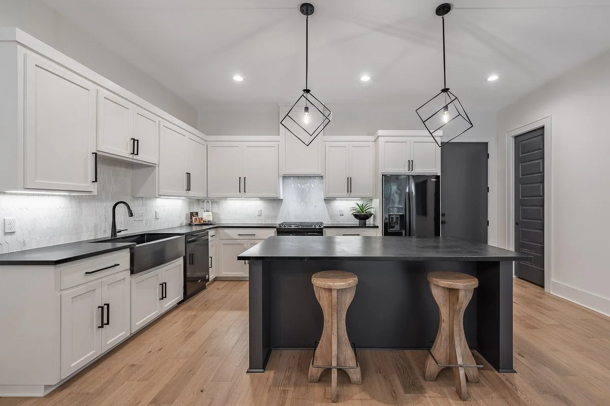 Real Estate photo of a modern kitchen with white cabinets, black countertops, black appliances, a black island with two wooden bar stools, and two pendant lights hanging above the island.