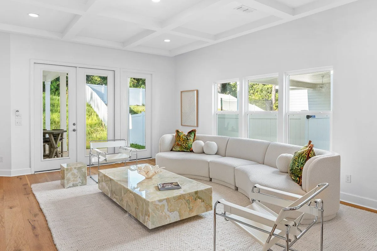 Real Estate photo of a bright living room with white walls, a beige sectional sofa with decorative pillows, a marble coffee table, and large windows and glass doors showing a grassy backyard. Minimalist decor with a white rug on wooden floors.