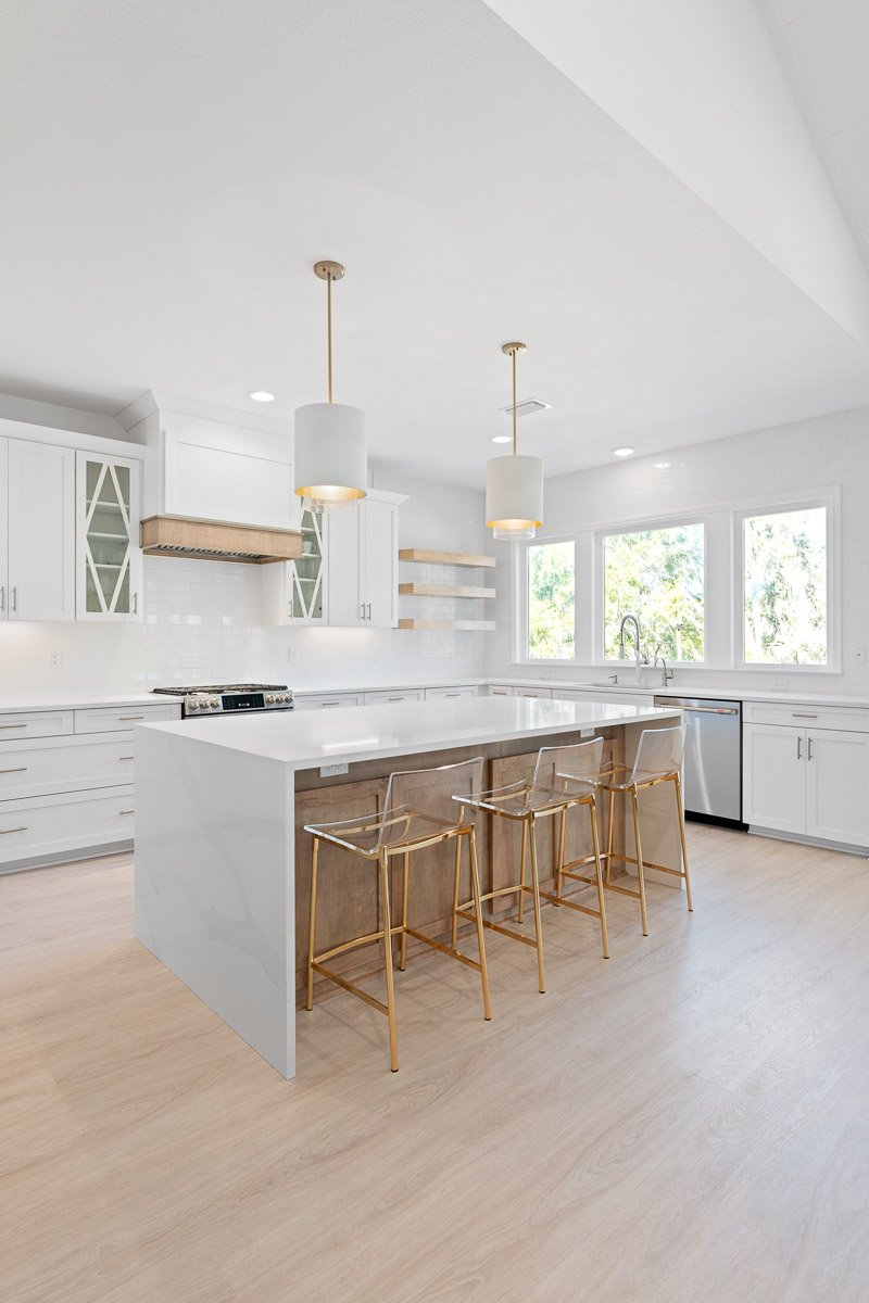 Real Estate photo of a bright, modern kitchen with white cabinetry, a large central island, gold bar stools, pendant lighting, and a window view of greenery.