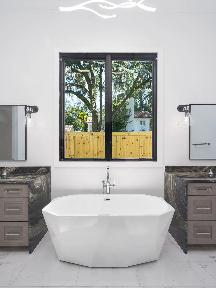 Modern bathroom with a freestanding bathtub between two vanities with dark marble countertops, large window showing trees and a wooden fence outside, white walls, and minimalistic lighting.