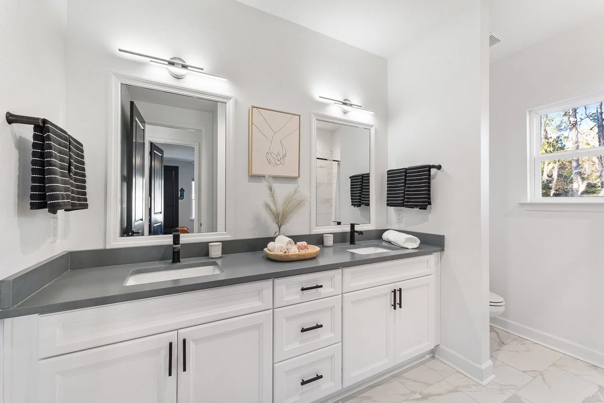 Real Estate photo of a modern white bathroom vanity with two sinks and black fixtures, gray countertops, two mirrors, black towels, decorative items, and a window with a view of trees.