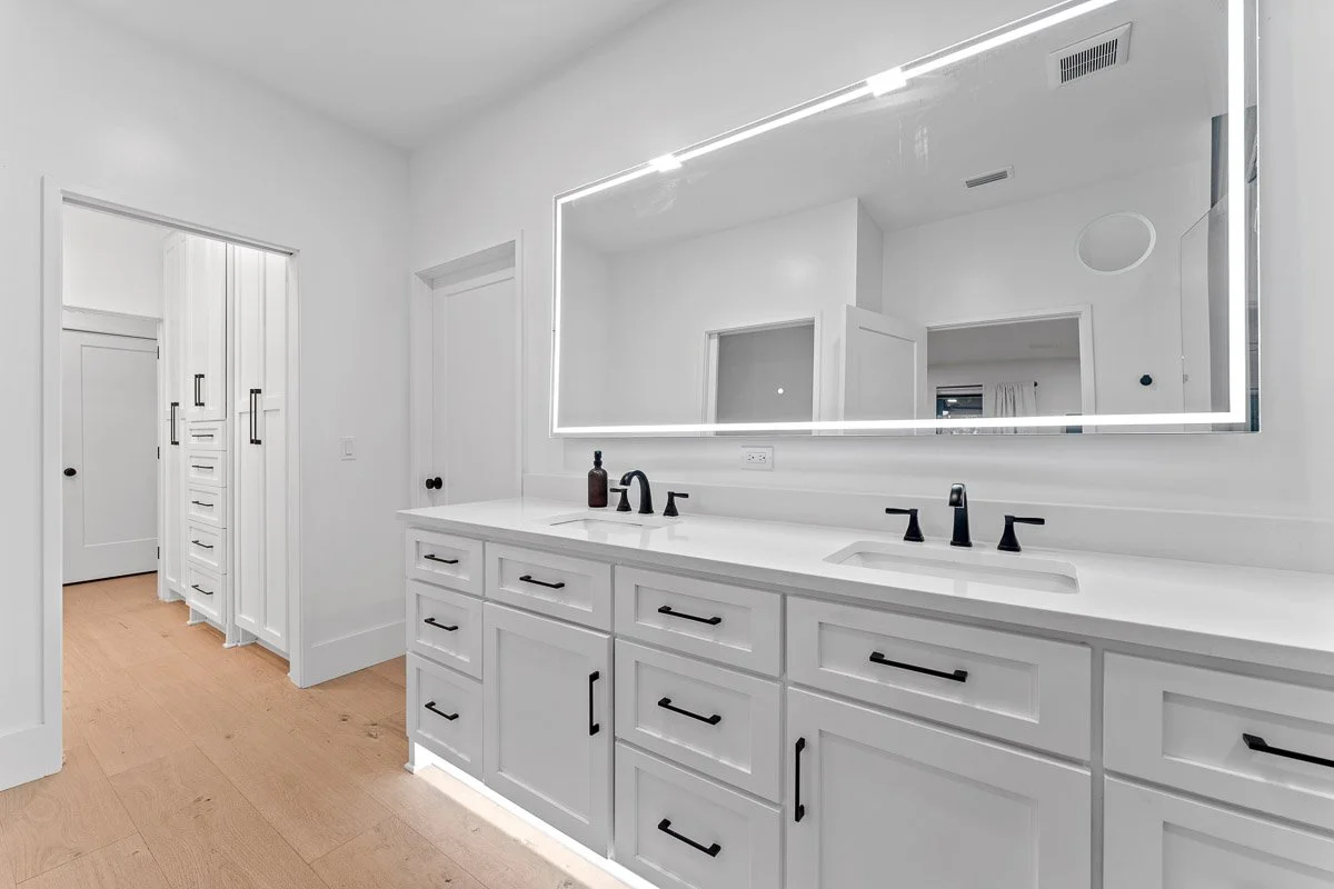 Real Estate photo of a modern white bathroom with a large illuminated mirror, black faucets, white cabinetry, and a light wood floor.