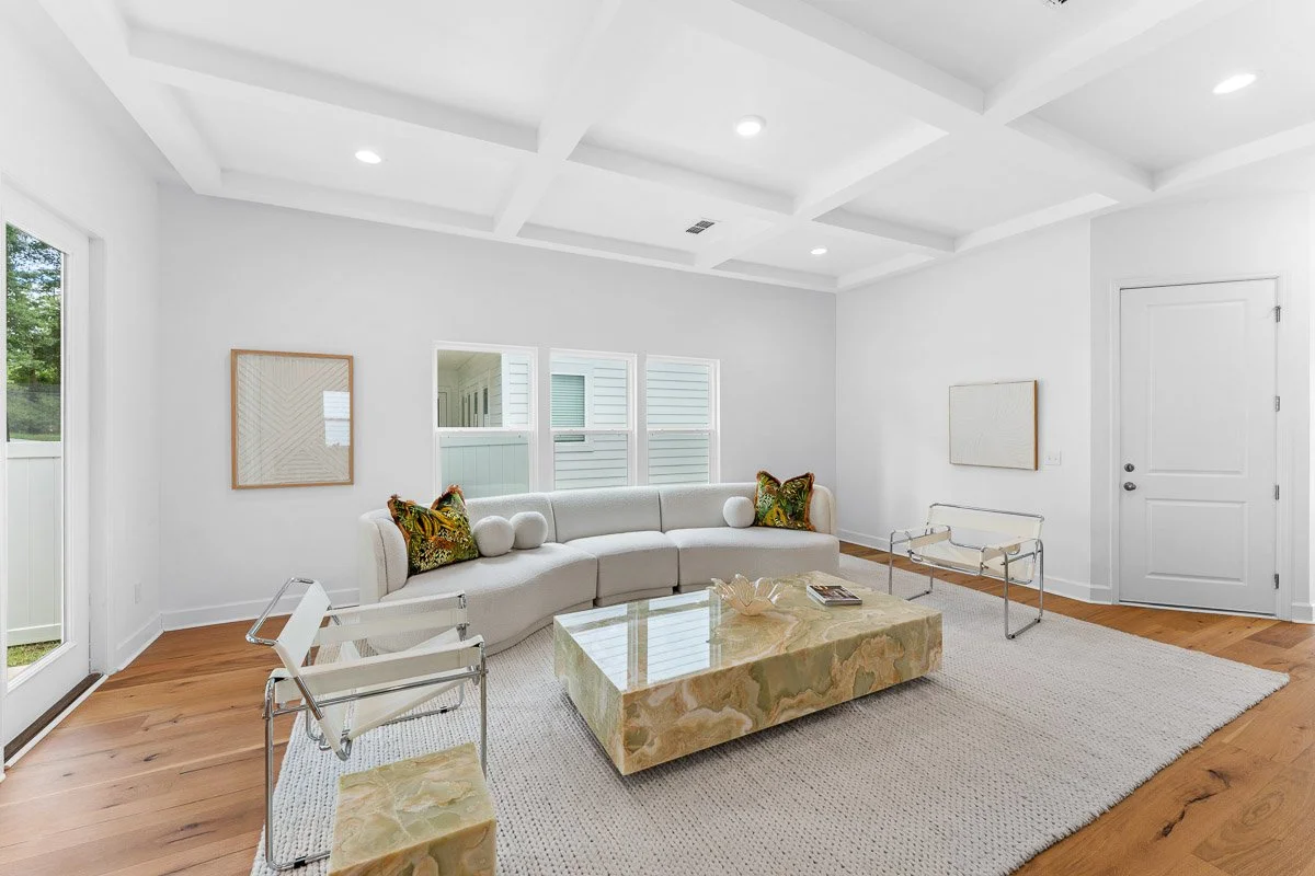 Real Estate photo of a minimalist living room with white walls, a curved white sofa with decorative pillows, a large beige marble coffee table, two white modern chairs, and a white area rug.