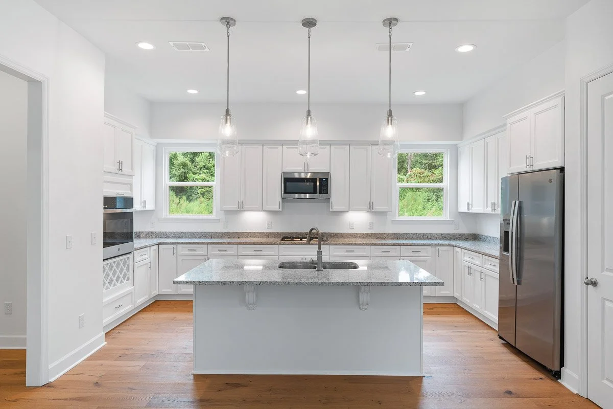 Real Estate photo of a bright, modern kitchen with white cabinets, granite countertops, a kitchen island with a sink, stainless steel appliances including a refrigerator and oven, and three pendant lights hanging above the island.