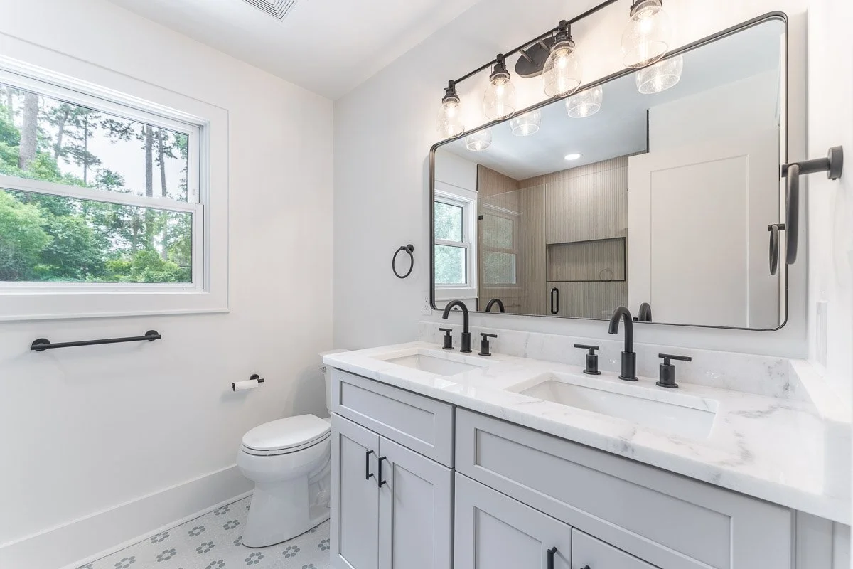 Real Estate photo of a newly renovated modern bathroom with white walls, a double sink vanity with black fixtures, a large mirror, and a window showing greenery outside.