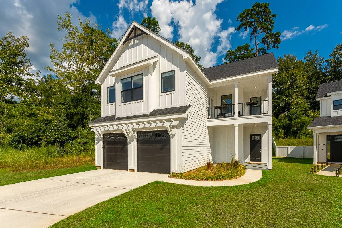 Real Estate photo of a modern two-story white house with black garage doors, black roof, and a small balcony, surrounded by green trees and grass.