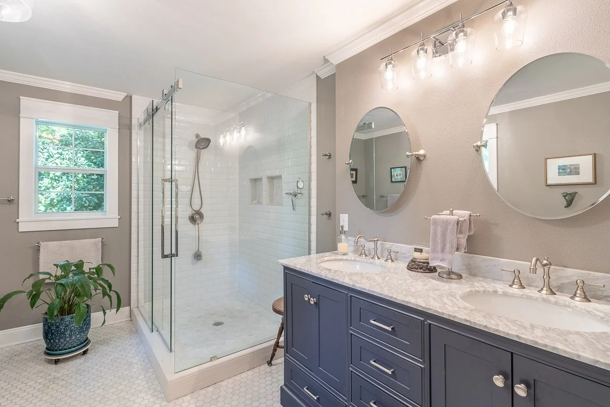 Real Estate photo of a modern bathroom with a glass-enclosed shower, a double sink vanity with a marble countertop, and round mirrors. There is a window with greenery outside, and a potted plant on the floor.