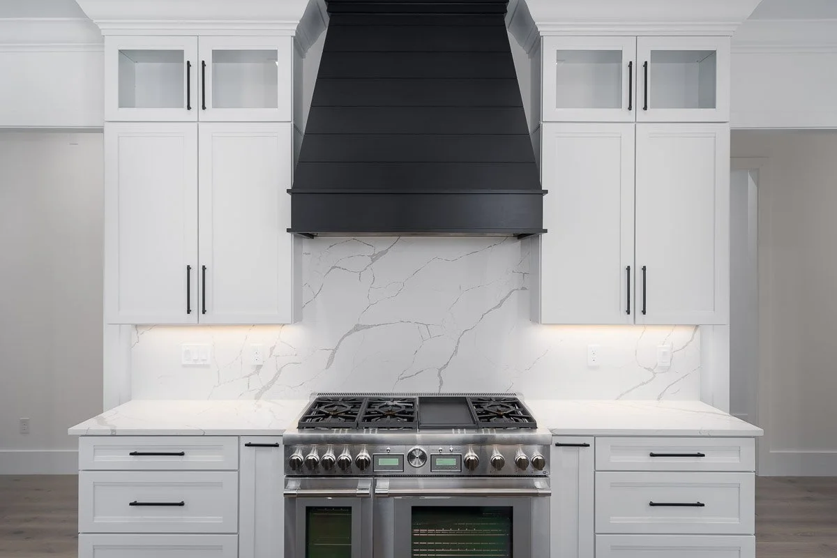 Modern kitchen with white cabinets, black handles, marble backsplash, and a stainless steel gas stove with knobs and oven, black range hood above.