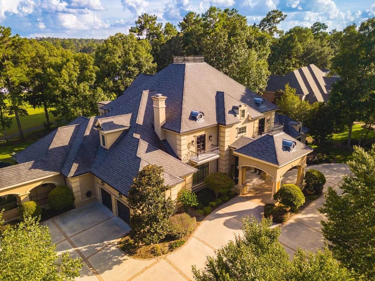Aerial Real Estate photo of view of a large, elegant house with multiple grey shingle roofs, surrounded by green trees and a landscaped yard with bushes and a driveway.