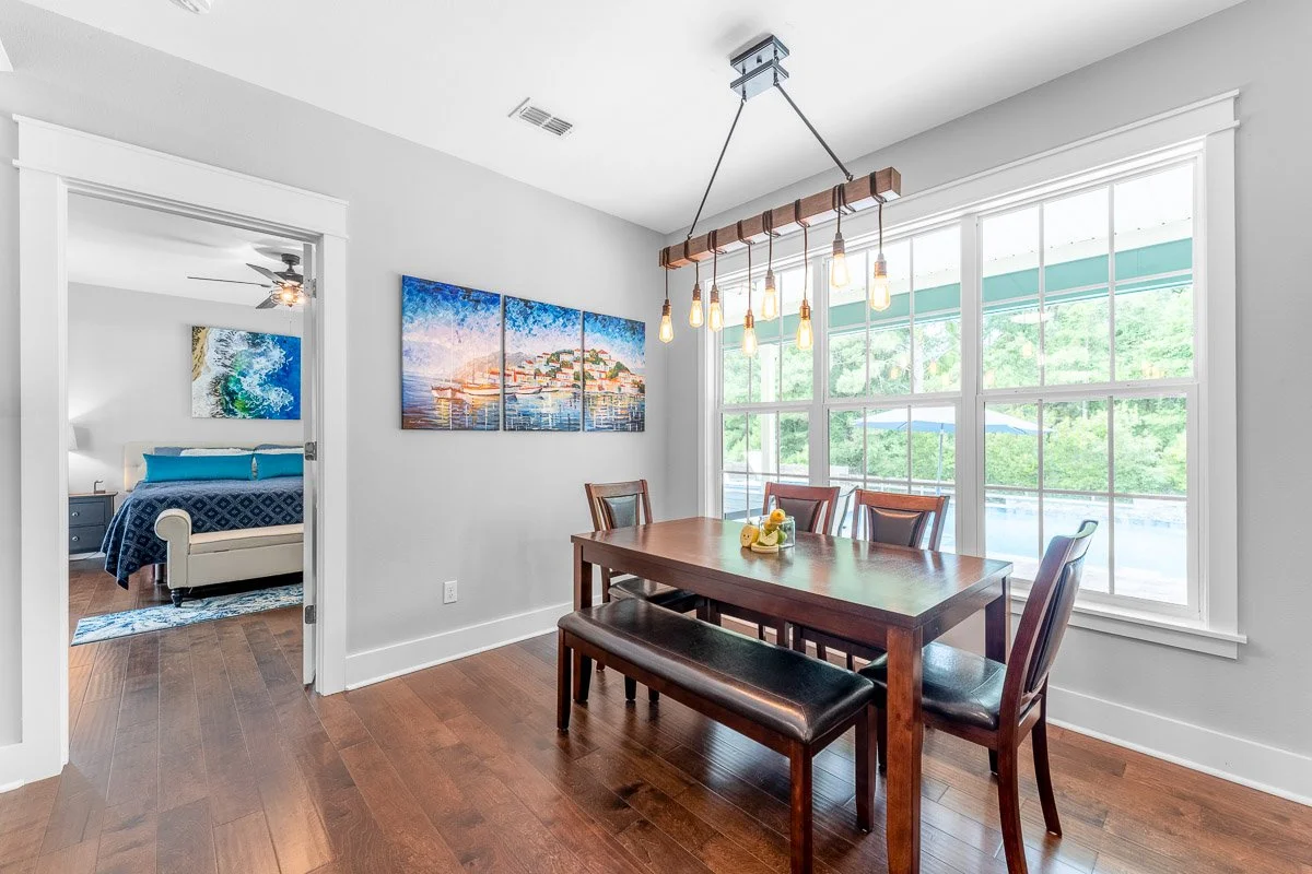 Real Estate photo of a dining room with wooden table, four chairs, large window, and modern light fixture, adjacent to bedroom with art on wall.