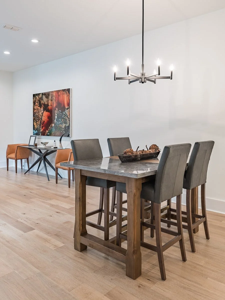 Real Estate photo of a modern dining room with a wooden table and four upholstered chairs, a black chandelier above the table, and a wall with a colorful abstract painting. In the background, a small side table with two orange chairs and decorative i
