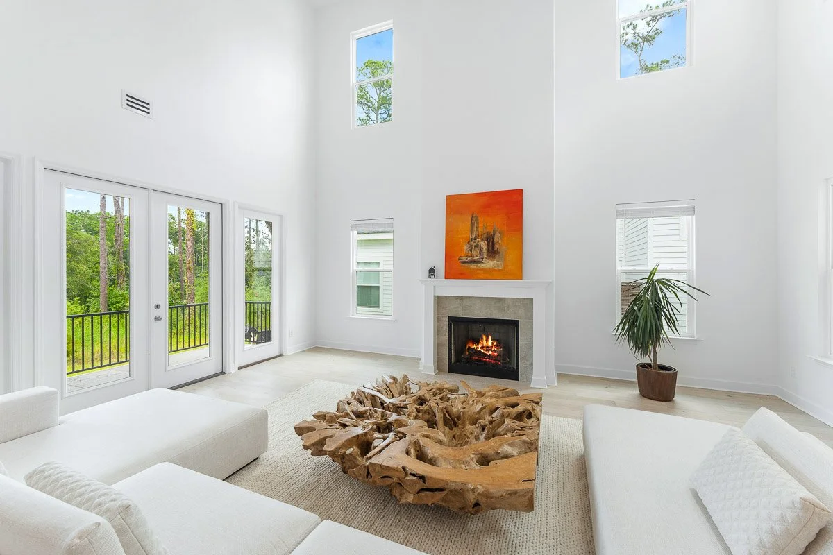 Real Estate photo of a living room with white walls, high ceiling, large windows, French doors leading outside, a fireplace with an orange painting above, a potted plant, and a unique wooden coffee table.